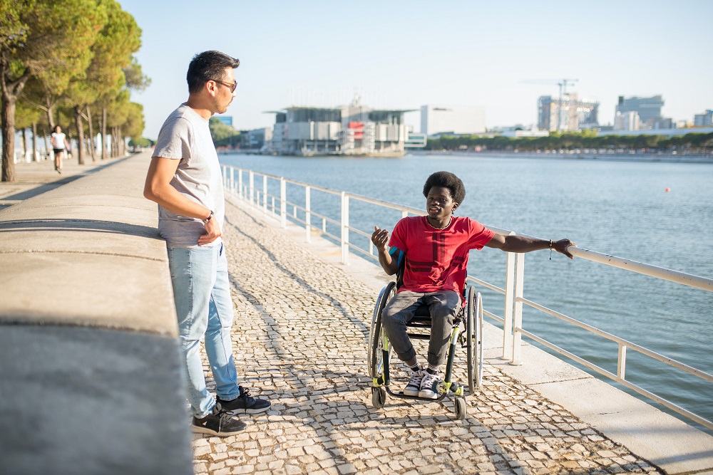 young-man-on-wheelchair-holding-rail-talking-to-man-on-river-bank